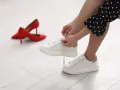Kobieta sznuruje białe trampki, które wydają się trochę za duże. Obok leżą czerwone szpilki.