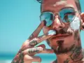 Mężczyzna z tatuażami i monroe piercingiem nakłada krem z filtrem na twarz, odbijając się w okularach.