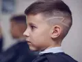 Chłopiec z podgolonymi bokami i wzorem na włosach, o pociągłej twarzy, czeka na zakończenie fryzury.