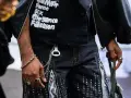 Mężczyzna w czarnej kurtce i spodniach z zamkami błyskawicznymi, ozdobionych cyfrowym wzorem. Stylizacja nawiązuje do **stylu opium**, z widocznym naszyjnikiem i pierścionkami.