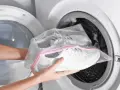 Ręce wkładają białe buty sportowe w siatkowej torbie do pralki. Pranie butów w pralce to szybki sposób na odświeżenie ulubionych sneakersów.