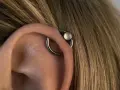 Oryginalny kolczyk orbital piercing z białym kamieniem na uchu, otoczony blond włosami.