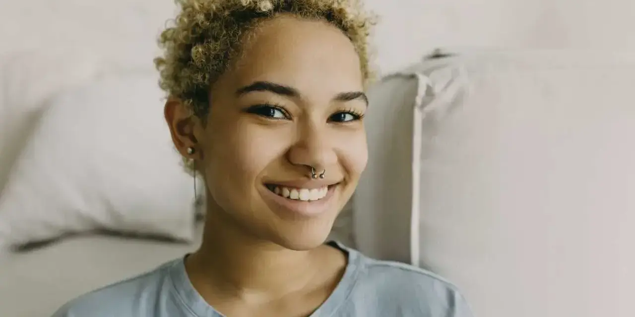 Uśmiechnięta młoda kobieta z kręconymi blond włosami i kolczykiem w nosie (septum piercing).
