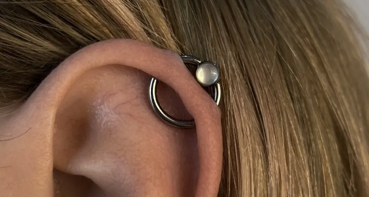 Oryginalny kolczyk orbital piercing z białym kamieniem na uchu, otoczony blond włosami.