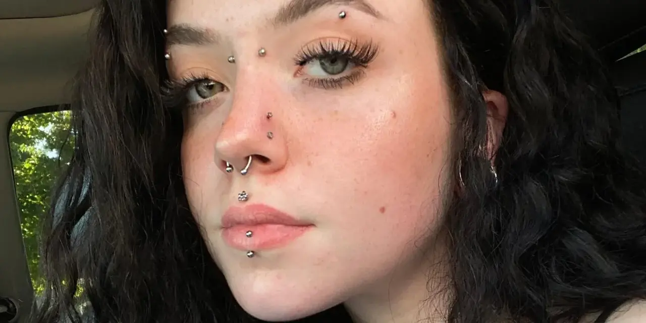 Dziewczyna z licznymi kolczykami: w nosie, ustach, brodzie i na czole. Wyróżnia się anti eyebrow piercing.