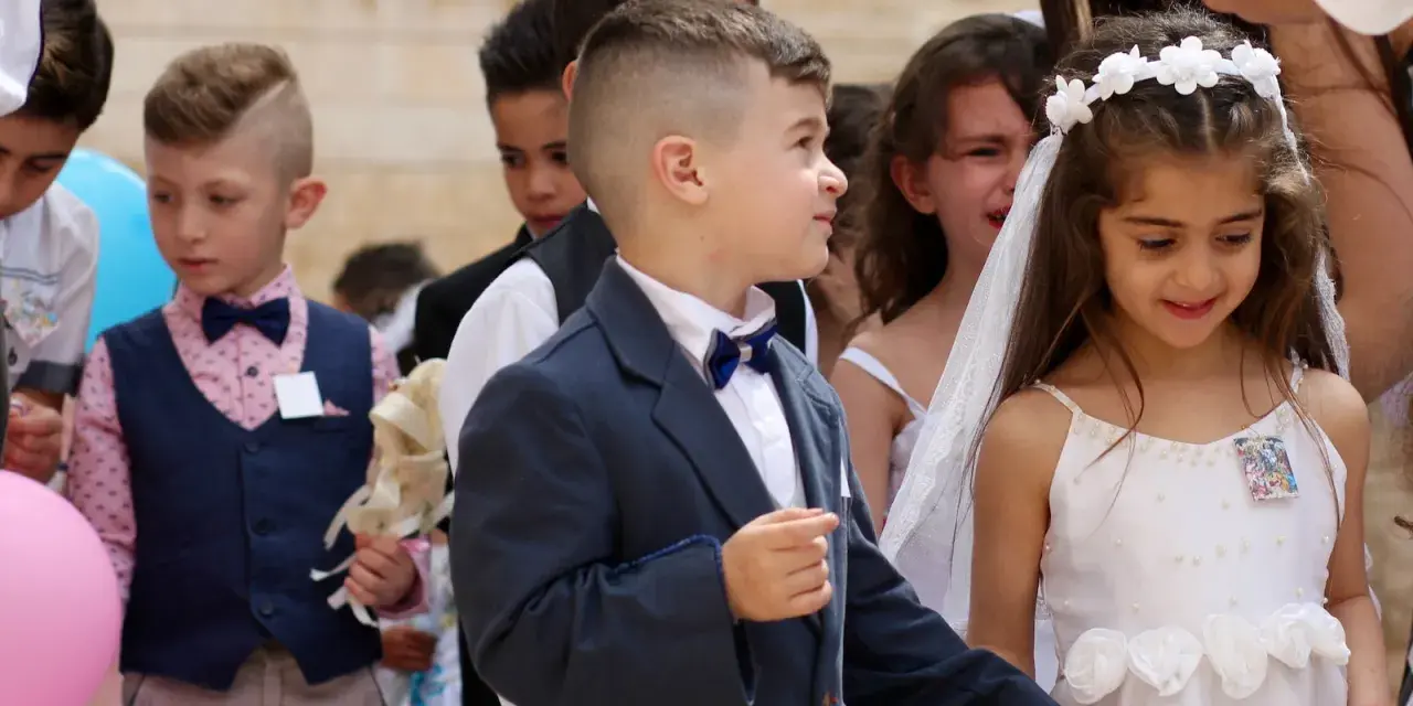 Chłopiec w eleganckim garniturze, z kokardką, wygląda jak z obrazka pin-up. Obok niego dziewczynka w białej sukience.