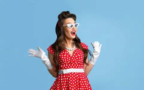 Kobieta w czerwonej sukience w grochy, białych rękawiczkach i okularach w stylu pin up, z uniesionymi rękami, wyraża radość.