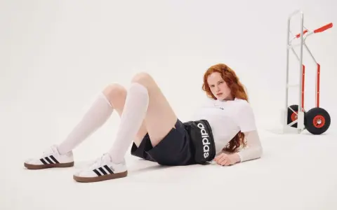 Rudowłosa modelka w białej koszulce i czarnych spodenkach z logo Adidas leży na podłodze. Czy Adidas ma zawyżoną rozmiarówkę?