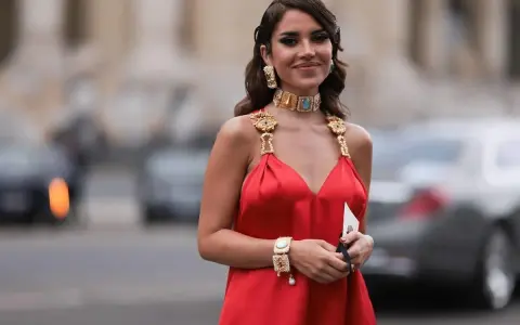 Elegancka kobieta w czerwonej sukience, prezentująca bogatą, złotą biżuterię: naszyjnik, kolczyki i bransoletkę. Idealna inspiracja, jaka biżuteria do czerwonej sukienki.