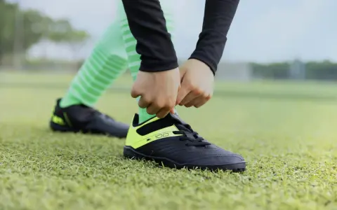 Dłonie wiążą sznurówki czarnych butów sportowych z neonowożółtymi wstawkami na zielonej murawie. To pierwszy krok, by dowiedzieć się, jak rozciągnąć buty sportowe.