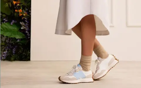 Elegancka sukienka i sportowe buty tworzą stylowy kontrast. Nogi w beżowych skarpetkach i białych sneakersach z błękitnym logo.