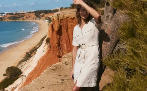 Kobieta w lnianej szmizjerce z paskiem stoi przy drzewie na klifie nad plażą.