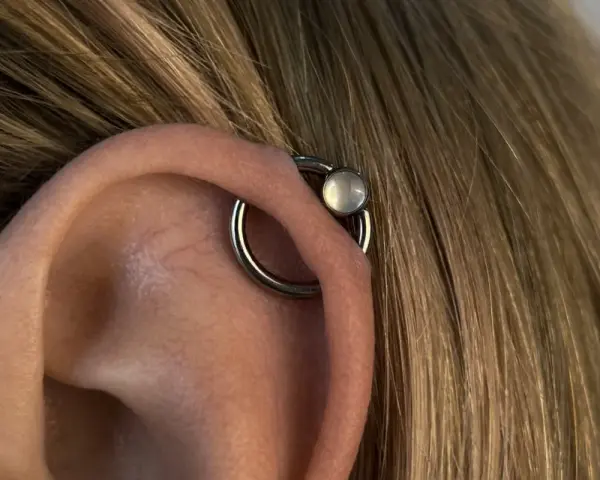 Oryginalny kolczyk orbital piercing z białym kamieniem na uchu, otoczony blond włosami.