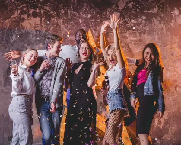 Grupa młodych ludzi świętuje, sypią się konfetti. Inspiracja, jak się ubrać do klubu: sukienka, jeansy, topy, marynarka.
