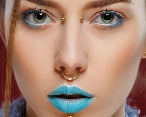 Kobieta z błękitnymi ustami i złotym septum. Jej oczy podkreślone są niebieskim cieniem, a między brwiami widoczny jest bridge piercing.