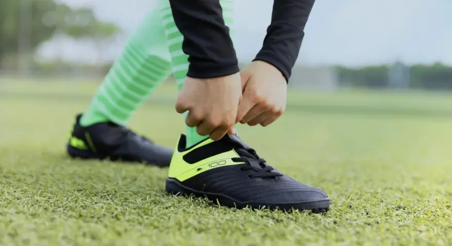 Dłonie wiążą sznurówki czarnych butów sportowych z neonowożółtymi wstawkami na zielonej murawie. To pierwszy krok, by dowiedzieć się, jak rozciągnąć buty sportowe.