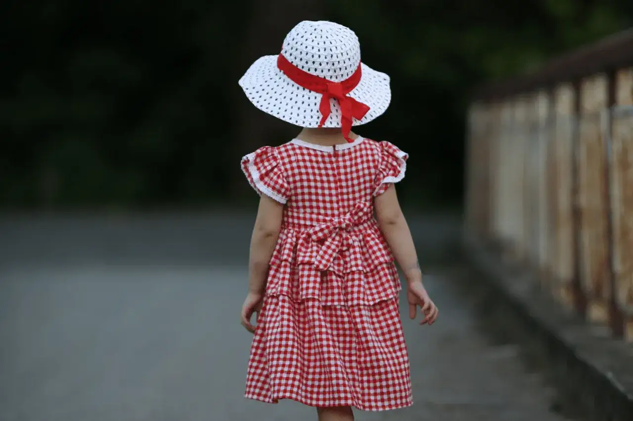 Dziewczynka w sukience w kratkę i kapeluszu z czerwoną wstążką, jak z obrazka pin-up, spaceruje ścieżką.